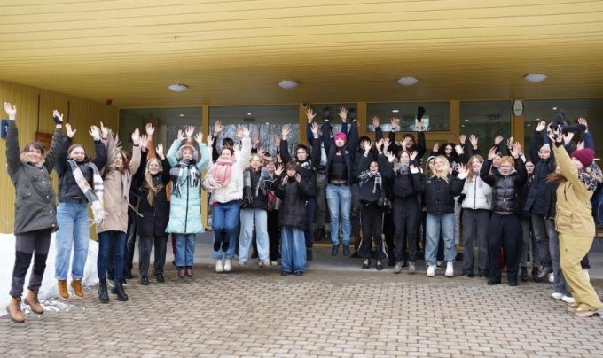 Skolēnu grupa stāv pie ēkas ieejas un ar paceltām rokām priecīgi sveicina