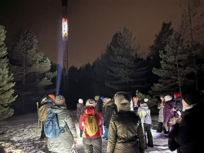 Pārgājiena dalībnieki mežā tumsā vēro izgaismoto uguns apsardzības torni