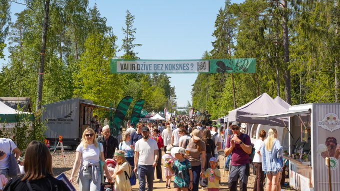 Saulainā laikā cilvēki uz grants ceļa pastaigas, apkārt izvietoti pasākuma stendi un teltis