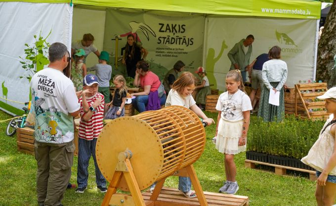 Maza meitene miniLAMPA zonā ar roku griež čiekuru žāvēšanas seno iekārtu, kas gatavota no koka, apkārt stāv vēl vairāki bērni