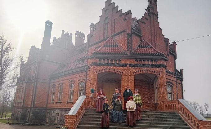 Sievie&scaron;u grupa tautastērpos stāv uz kāpnēm pie Jaunmoku pils - vēsturiskas sarkano ķieģeļu ēkas
