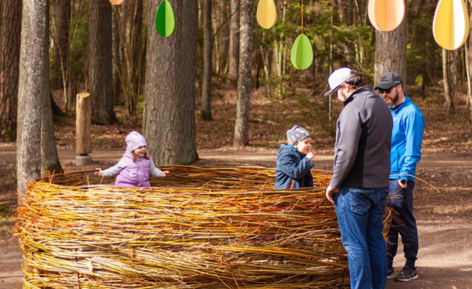 Lieldienu aktivitātes &ndash; divi bērni rotaļājas lielā no klūgām veidotā ligzdā, pie kuras stāv viņu tēti. Virs cilvēkiem iekarinātas krāsainas papīra olas