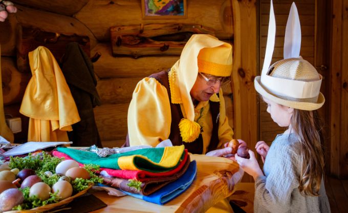 Lieldienu noskaņās Tērvetes rūķis dzeltenā cepurē ar lielu pušķi galā pēta krāsoto olu kopā ar mazu meiteni, kurai galvā cepure ar pielīmētām zaķa ausīm