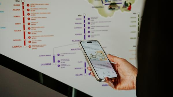 Vienā rokā tur telefonu ar kartes attēlu, fonā uzrakstu shēma