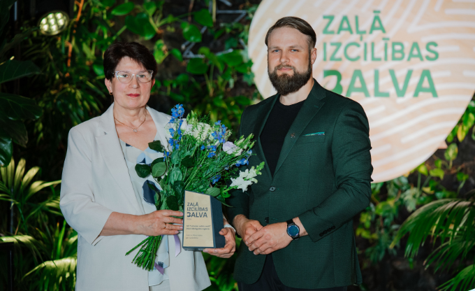 Divi cilvēki pasākumā pozē ar balvu un ziedu pušķi uz zaļa augu fona
