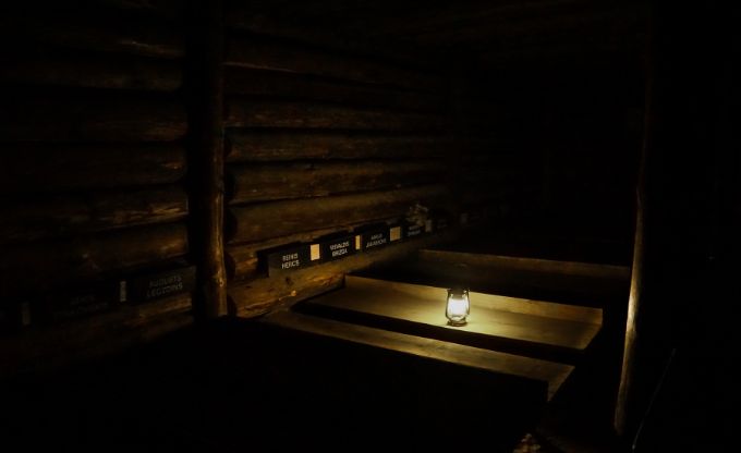 Tumšs koka bunkurs, kur uz koka galda centrā deg neliela laterna, izgaismojot apkārtni