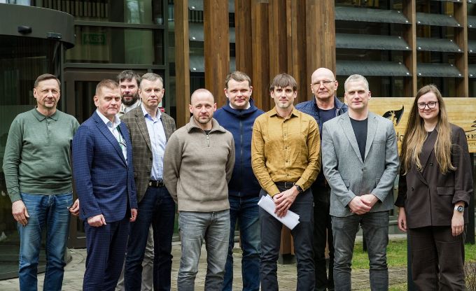 Grupa ar deviņiem cilvēkiem stāv ārā pie modernas koka un stikla ēkas ieejas, pozējot kopbildei