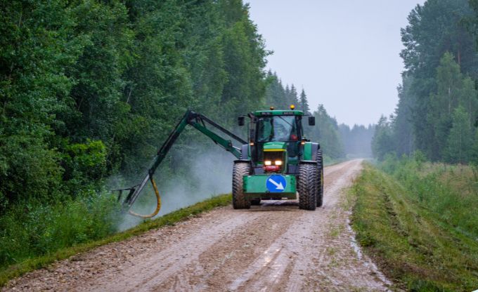 Ar piekabināmu pļaujmašīnu aprīkots lauksaimniecības traktors zaļā krāsā uz meža autoceļa veic atva''su pļaušanu ceļa grāvī, ir apmākusies vasaras diena