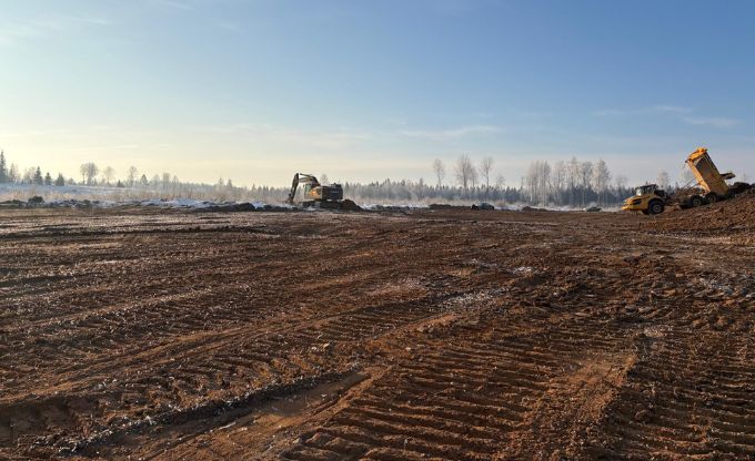 Plašā laukumā ar tehnikas atstātām pēdām ekskavators strādā tālumā, kur fonā redzams neliels sniegs