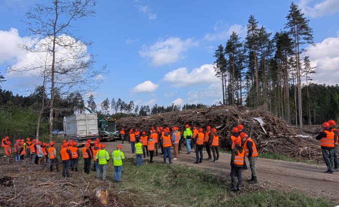 Cilvēki oranžās vestēs un ķiverēs pulcējas mežā pie šķeldas kaudzes un šķeldotāja