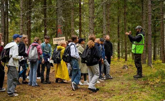 Skolēnu grupa stāv mežā kopā ar LVM darbinieku